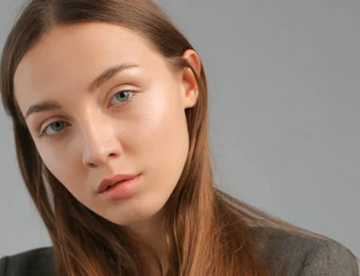 A young woman with long, straight, light brown hair wearing a grey blazer, posing against a neutral grey background. Her expression is calm and thoughtful, and her hair falls naturally over her shoulders.