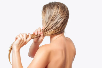 A woman with wet, blonde hair styled into a loose braid, seen from behind against a white background. Her bare shoulders and back are visible, emphasizing the hair texture and styling process. 