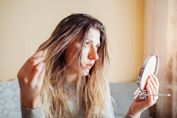 A young woman looking at her greasy hair in a handheld mirror.